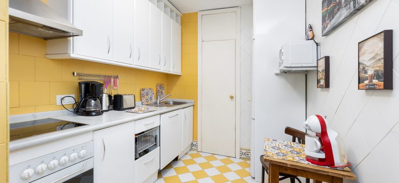 Cocina, tonos amarillos, mesa auxiliar, cuadros de cafés con leche, armarios, horno y lavavajillas blancos, cafetera y tostadora sobre la encimera