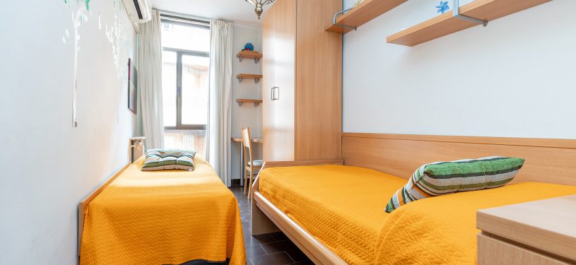 Habitación de dos camas individuales, tonos amarillo/naranja, paredes blancas, con armario y escritorio