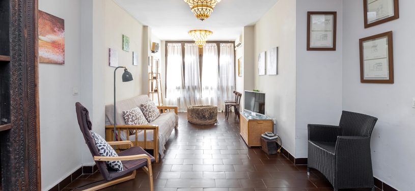Salón con amplio ventanal, cortinas blancas, sofá, televisor enfrente, estanterías de madera, sillas y espejo